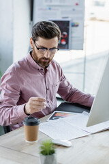 Businessman working with papers and computer near coffee to go in office.