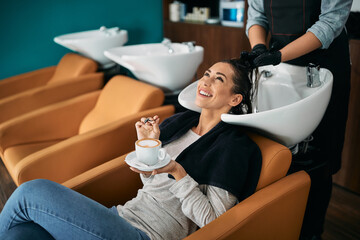 Happy woman enjoys in cup of coffee during hair wash at hairdresser's.