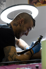 vertical portrait of a smiling crested tattooist tattooing a client's leg in his tattoo studio