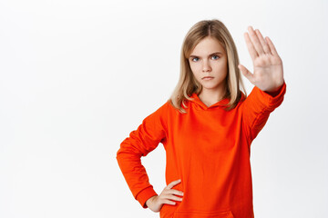 Stop. Little girl looks serious, extends one arm to say no, rejecting, prohibit action, forbid smth, standing in red hoodie over white background