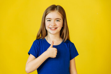 cute little girl gesturing thumbs up and looking at camera smiling to camera, yellow background. Children sincere recommendation. Like and approval. - obrazy, fototapety, plakaty