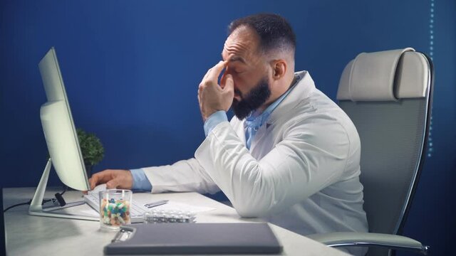 Tired Doctor In A Medical Office At A Work Desk In Front Of A Computer. Medicine And Health Care Concept