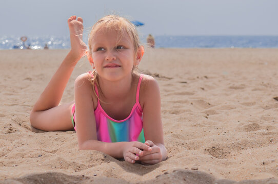 Little Girl, Blonde, 6 Years Old, Lies In A Bright Bikini On The Sand, On The Beach, By The Sea, A Child's Portrait Of A Sunbathing Girl On The Sea Coast