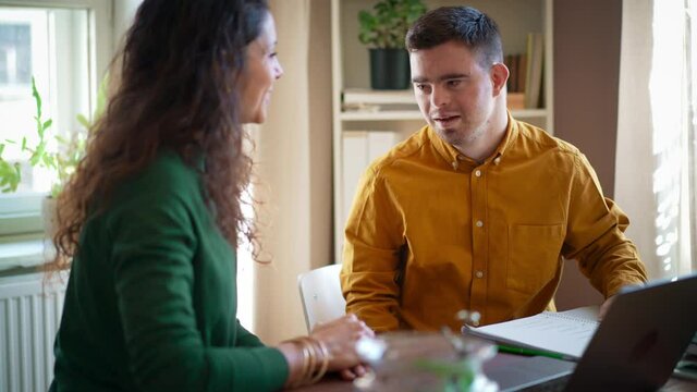 Young man with Down syndrome with his tutor studying and using laptop indoors at home, homeschooling.