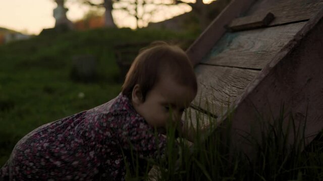 Authentic cute little infant baby girl in dress try to climb the wooden stairs at spring sunset. playful child crawley lawn on nature during sun rise. Childhood, parenthood, helplessness concept