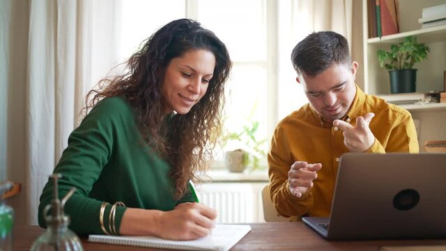 Young man with Down syndrome with his tutor studying and using laptop indoors at home, homeschooling.