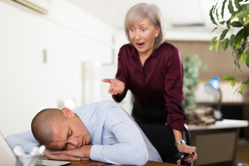 Man office worker sleeping on table during work day. His chief senior woman standing next to him and shouting at him.