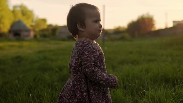 Authentic cute little infant baby girl in dress walking in park on tall grass at spring sunset. playful child crawley lawn on nature during sun rise. Childhood, parenthood, family, lifestyle concept