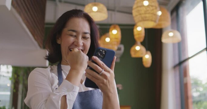 Asia mature adult woman people wear apron small cafe receive e-mail text good news SME lending service solution win seller store in social media app. Shock smile laugh face in sale order reopen shop.