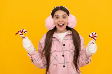 amazed kid in earmuffs and mittens hold candy stick on yellow background, new year