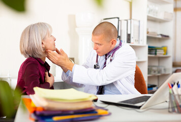 Senior woman visiting qualified doctor in medical office with complaining of sore throat. Focused therapist examining female patient and prescribing treatments while sitting at table - obrazy, fototapety, plakaty