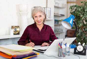 Portrait of confident smiling elderly female office employee during daily work with laptop and documents..
