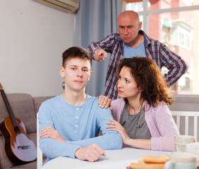 Frowning teen boy sitting at home while mother and father berating him ..