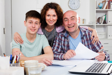 Portrait of happy friendly family of three with teenage son working with papers and laptop at home