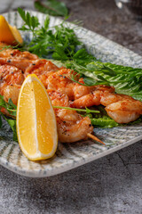 Grilled king prawns with lemon and herbs on a plate against a gray stone table, fish kebab