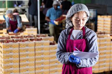 Asian woman supervisor with documents controlling quality of cherry sorting at fruit warehouse