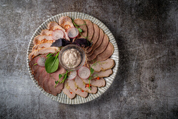 An appetizer in a restaurant, assorted meats. Sliced meat and sausage in layers on a plate with greens against a gray stone table 