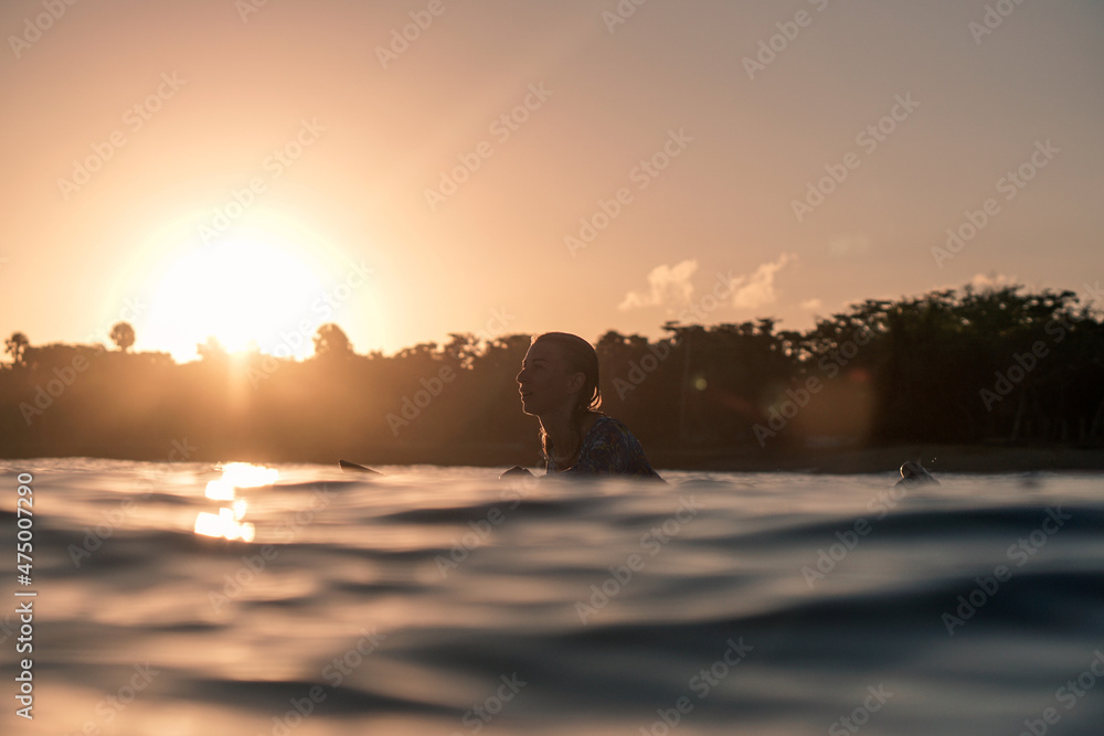 Wall mural Portrait of blond surfer girl on white surf board in blue ocean pictured from the water at golden sunrise time in Encuentro beach