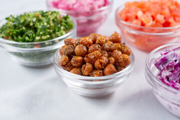 A view of a glass condiment bowl of deep fried garbanzo beans, among a variety of other chopped ingredients.