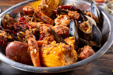 A view of a big silver bowl full of seafood boil, featuring mussels, clams, crawfish, shrimp, and a corn cob.