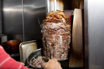 A view of an employee carving beef shawarma from a rotisserie spit machine, in a restaurant kitchen setting.