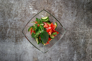 Summer salad of fresh cucumbers and tomatoes with herbs in a glass plate against a gray stone table