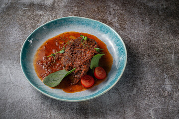 Beef stew with broth and herbs in a blue plate with spices and black pepper against a gray stone table .