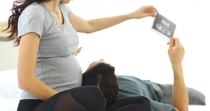 Pregnant woman and man looking at ultrasound photo while chilling on bed at home together
