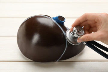 Doctor with stethoscope examining liver model at white wooden table, closeup