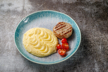 A mashed potato with a meat patty and fresh tomatoes on a blue plate against the background of a gray stone table.