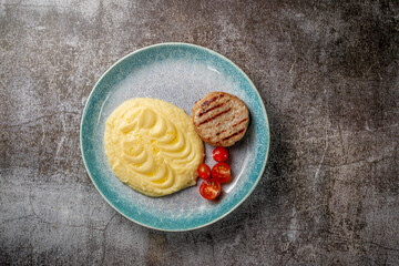 A mashed potato with a meat patty and fresh tomatoes on a blue plate against the background of a gray stone table.
