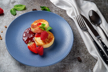 Fried pancakes with strawberry jam and raspberries and mint in a blue plate against a gray stone table. A sweet breakfast in the morning