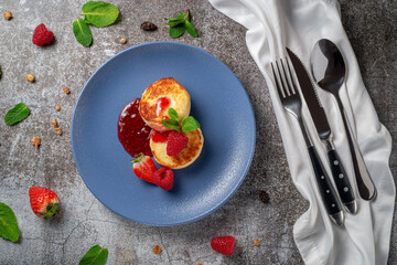 Fried pancakes with strawberry jam and raspberries and mint in a blue plate against a gray stone table. A sweet breakfast in the morning