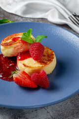 Fried pancakes with strawberry jam and raspberries and mint in a blue plate against a gray stone table. A sweet breakfast in the morning