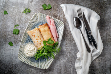 Pancakes filled with ham in a plate with parsley and dill greens and sour cream on a gray stone table, flatlay.