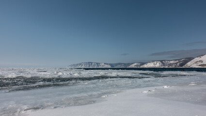 Fototapeta premium The ice on the river begins to melt. Footprints in the snow near the shore. In the distance - blue ice-free water. A mountain range against the azure sky. Siberia. Angara river