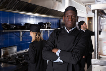 Confident African American chef of restaurant posing with arms crossed in kitchen on background with employees