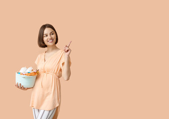 Young pregnant woman with baby clothes pointing at something on beige background