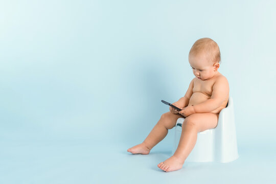 A newborn baby with a phone is sitting on a potty on a blue background. A child of 12-15 months is watching videos on a smartphone while sitting on a potty. Copy space