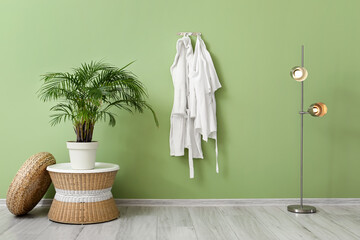 Houseplant with table, lamp and white bathrobes hanging on green wall - obrazy, fototapety, plakaty