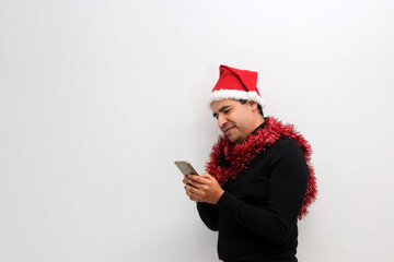 Latino adult man with Christmas hat and red garland as scarf uses his cell phone to call his family with happy video call and excited to celebrate Christmas
