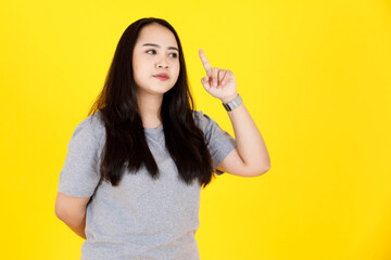 Portrait studio shot of Asian young chubby plump happy long black hair female model in gray casual tshirt outfit standing smiling pointing index finger up on blank copy space on yellow background