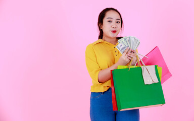 Fototapeta premium Portrait studio shot of Asian young rich wealthy female shopaholic shopper in casual outfit smiling holding hundred dollars fan shopping bags on pink background