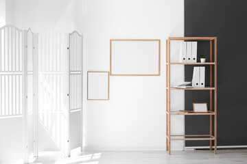 Folding screen, shelving unit with folders and blank frames hanging on light wall