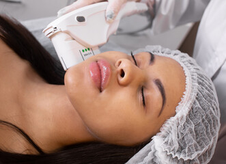 Obraz na płótnie Canvas Relaxed young black female client getting SMAS ultrasound face lifting massage with professional equipment in beauty salon.