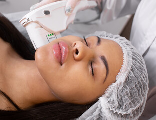 Obraz na płótnie Canvas Relaxed young black female client getting SMAS ultrasound face lifting massage with professional equipment in beauty salon.