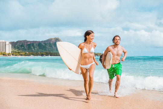 Hawaii surfing couple surfers in Waikiki beach, Honolulu Asian girl and Caucasian guy surfer carrying surfboards running out of water. Surf lifestyle Oahu island. USA travel. Fun vacation activity. - Powered by Adobe