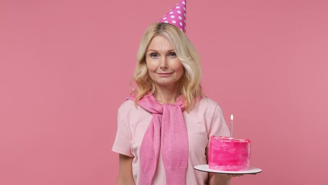 Fascinating jubilant exultant elderly blonde woman lady 40s years old wear t-shirt birthday hat hold cake with candle blow pipe celebrate isolated on plain pastel light pink background studio portrait