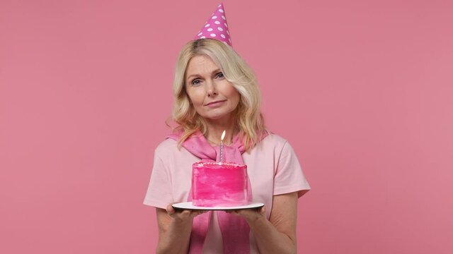 Sad distempered elderly blonde woman lady 40s years old wears t-shirt birthday hat look camera hold cake with candle sigh suspire sithe isolated on plain pastel light pink background studio portrait