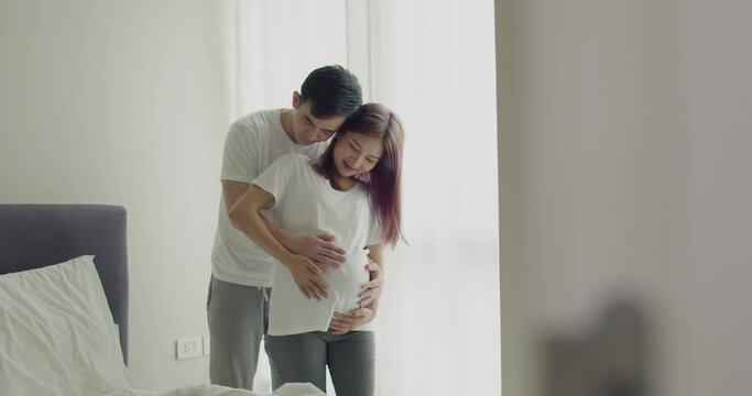 Happy Asian husband touching his wife's pregnant belly while standing beside a window in their beautiful bedroom.